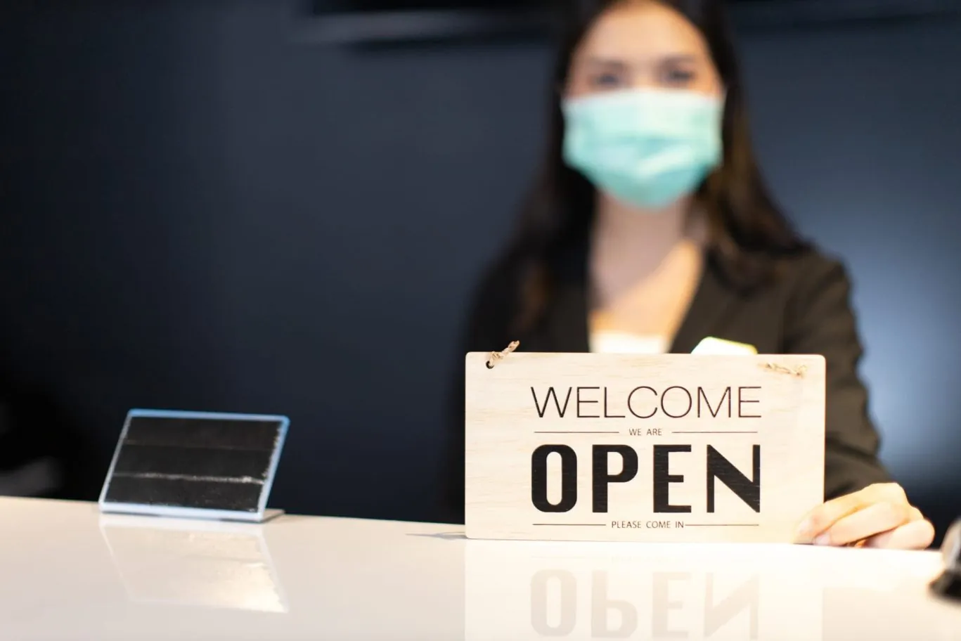 Woman at front reception desk with face covering holding a 'welcome, open' sign